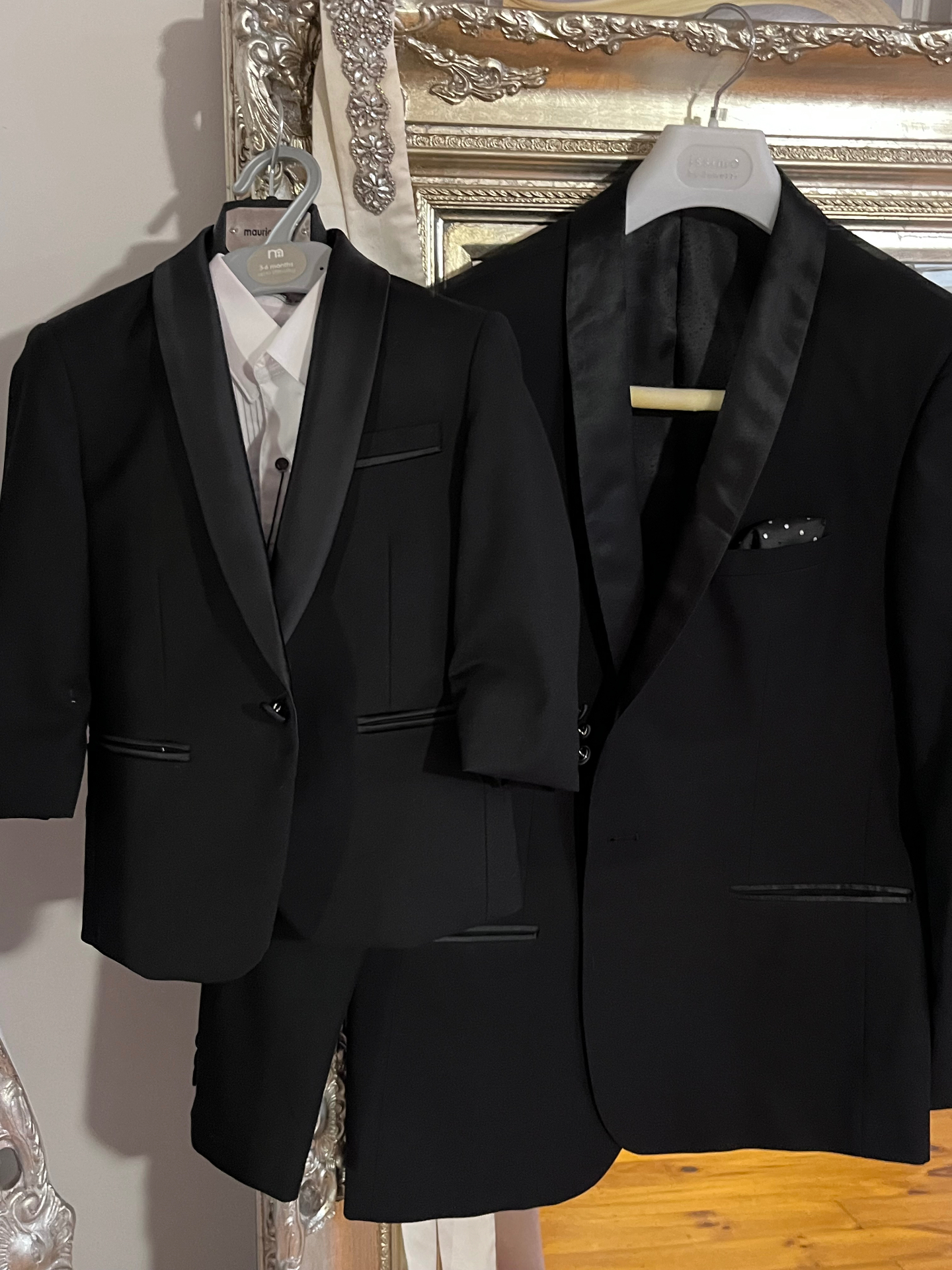 Two little page boy tuxedos with satin shawl lapels hanging in front of an ornate silver mirror