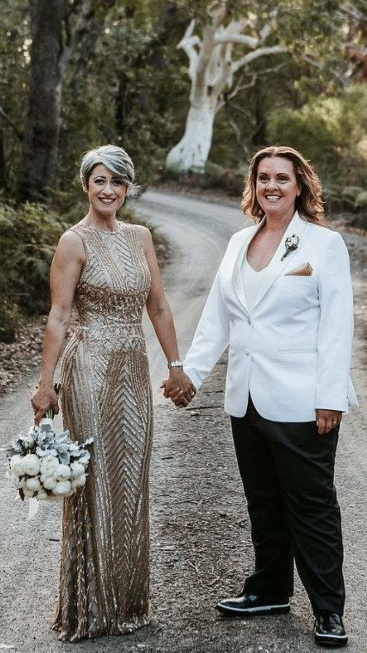 Two brides in formal wedding attire — one in a gold beaded column gown, the other in a tailored white suit jacket and black trousers — suit and formal gown alterations by Jacqui, NSW South Coast
