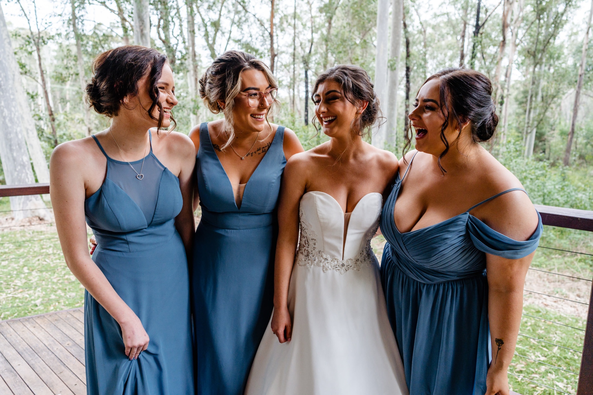Bridesmaids in mixed dusty blue gowns laughing with the bride, individually fitted by Jacqui