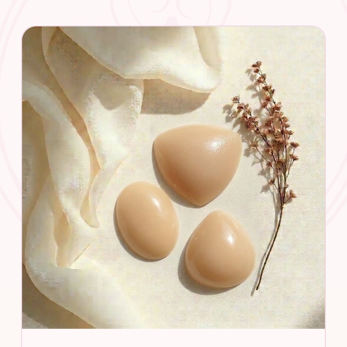Three beige Boomba silicone breast enhancers — triangle, oval and teardrop — styled on ivory linen with sheer silk and a dried blush floral sprig