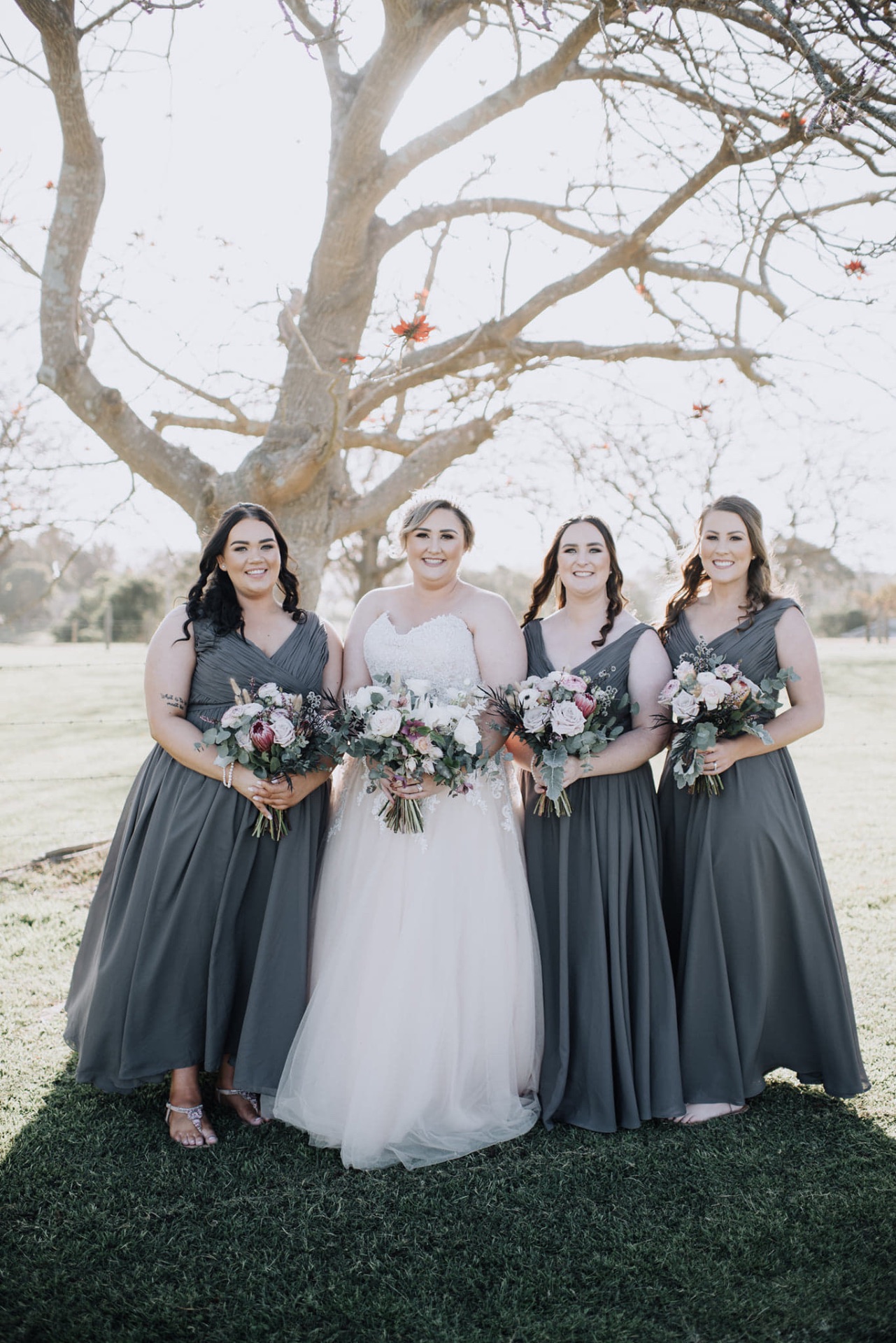 Bride with three bridesmaids in slate grey chiffon gowns under a tree, tailored on the NSW South Coast