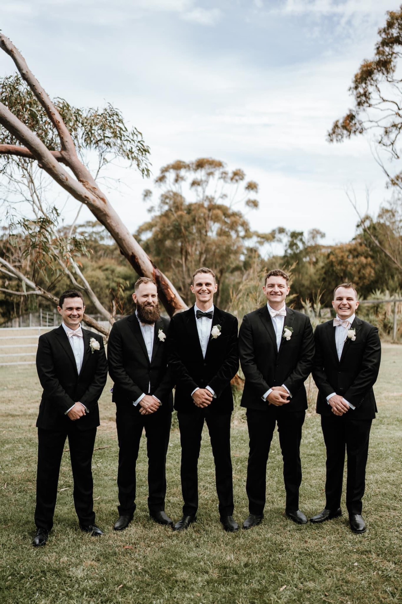 Groom and four groomsmen in black tuxedos with blush bow ties and buttonholes — coordinated groomsmen suit tailoring by Jacqui, NSW South Coast