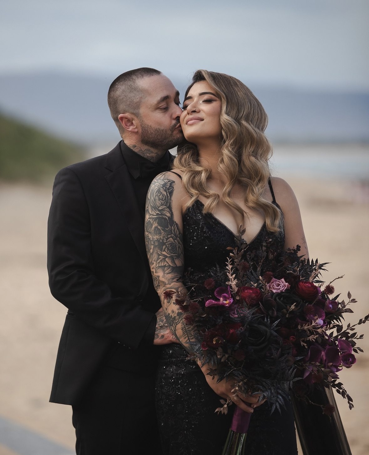 Bride in a black sequin gown holding a dark moody bouquet, kissed by groom in black tuxedo on a beach — evening gown alterations and tailoring by Jacqui